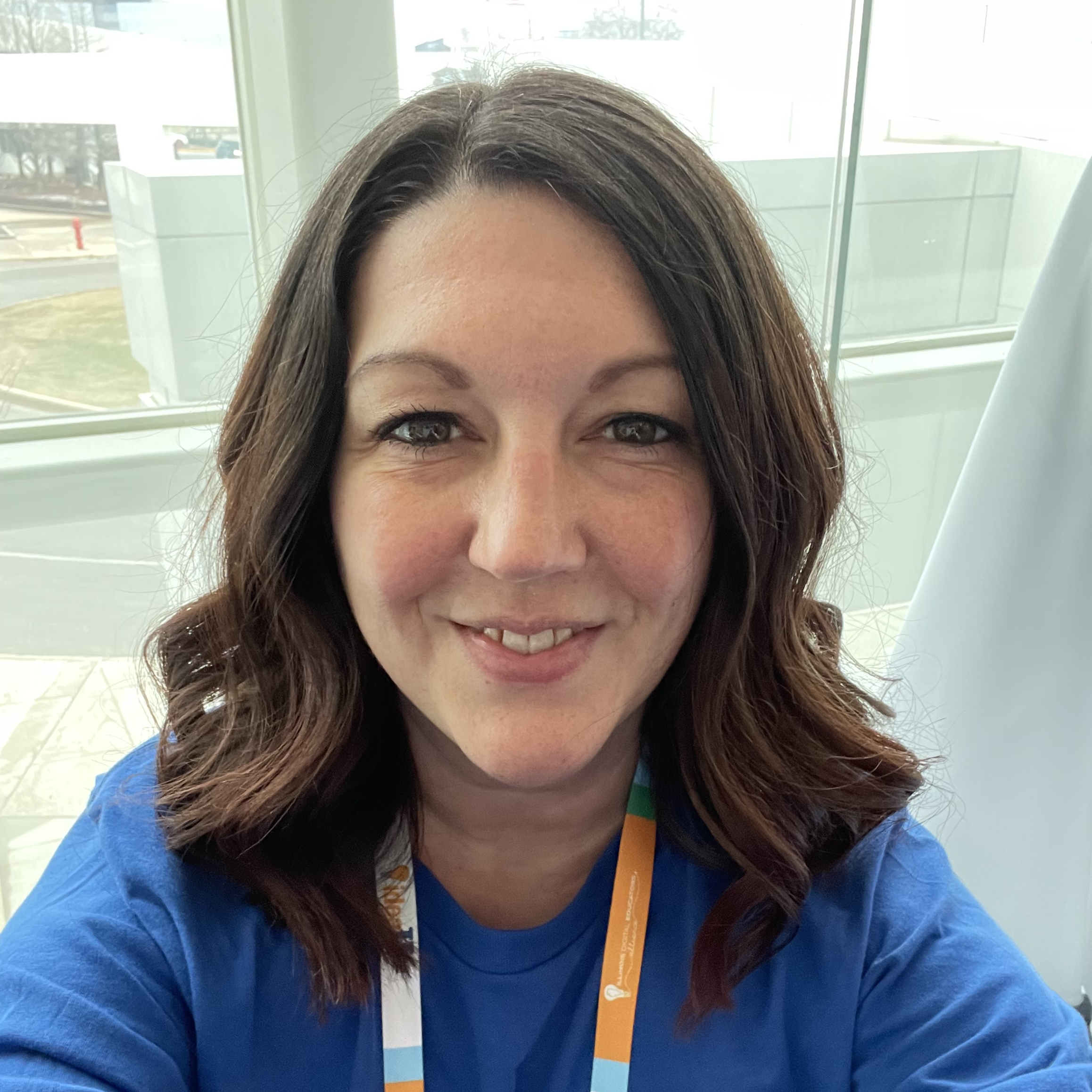 Woman with wavy brown hair wearing a blue shirt and lanyard, smiling at the camera indoors.