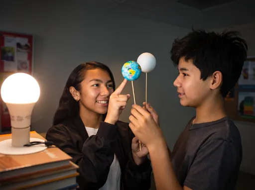 Two kids use models of Earth and Moon on sticks near a lamp to demonstrate orbit and light.