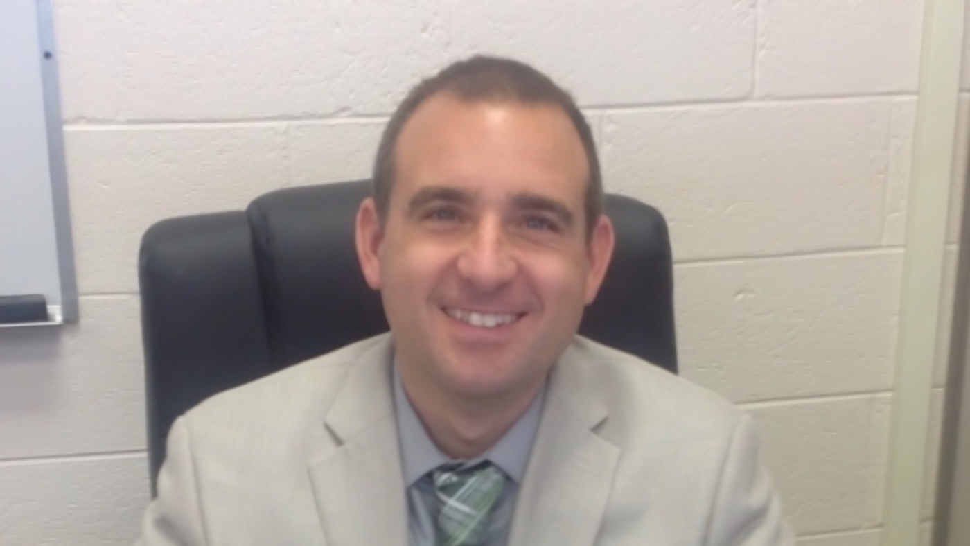 A man in a light-colored suit and tie smiles while sitting in an office chair against a white brick wall.