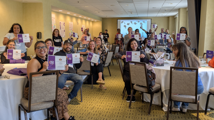 A group of people in a conference room smile and hold certificates while seated at round tables.