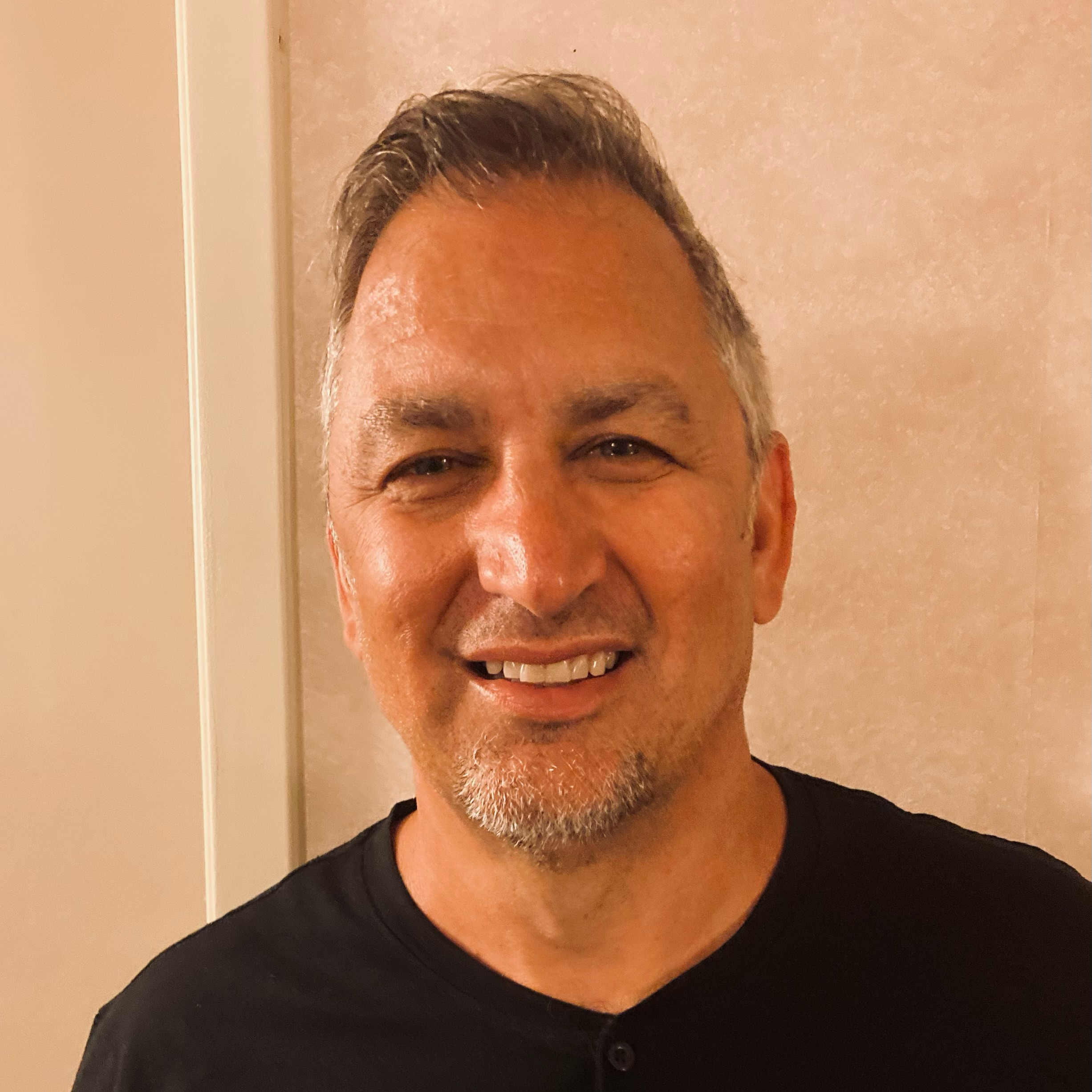 Smiling middle-aged man with short gray hair and a goatee, wearing a black shirt, stands in front of a beige wall.