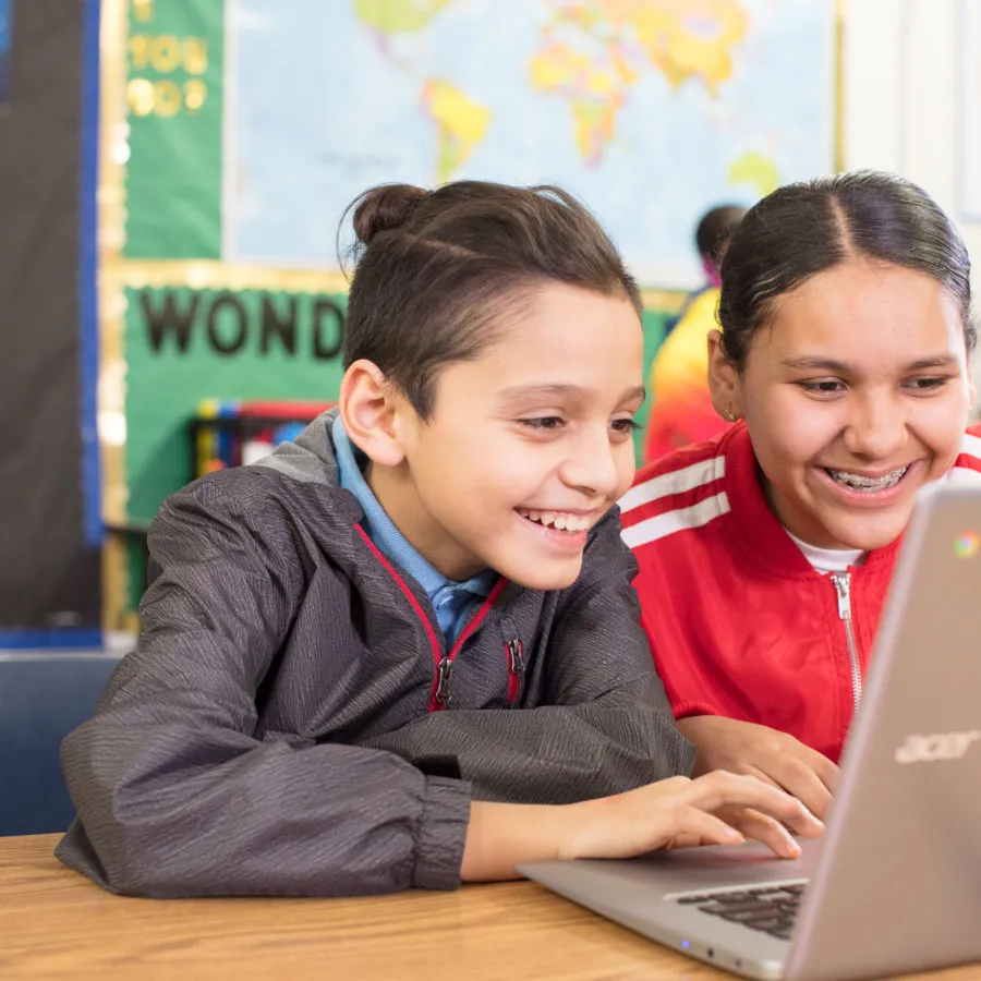 Two smiling children sit at a desk, looking at a laptop together in a colorful classroom.