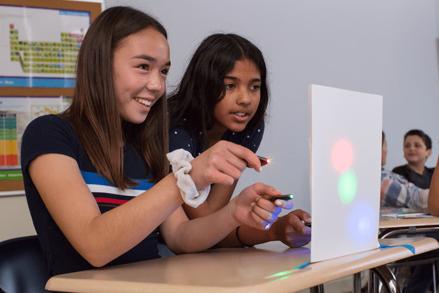 Two girls in a classroom shine colored lights onto a white board, creating colored circles, while smiling.