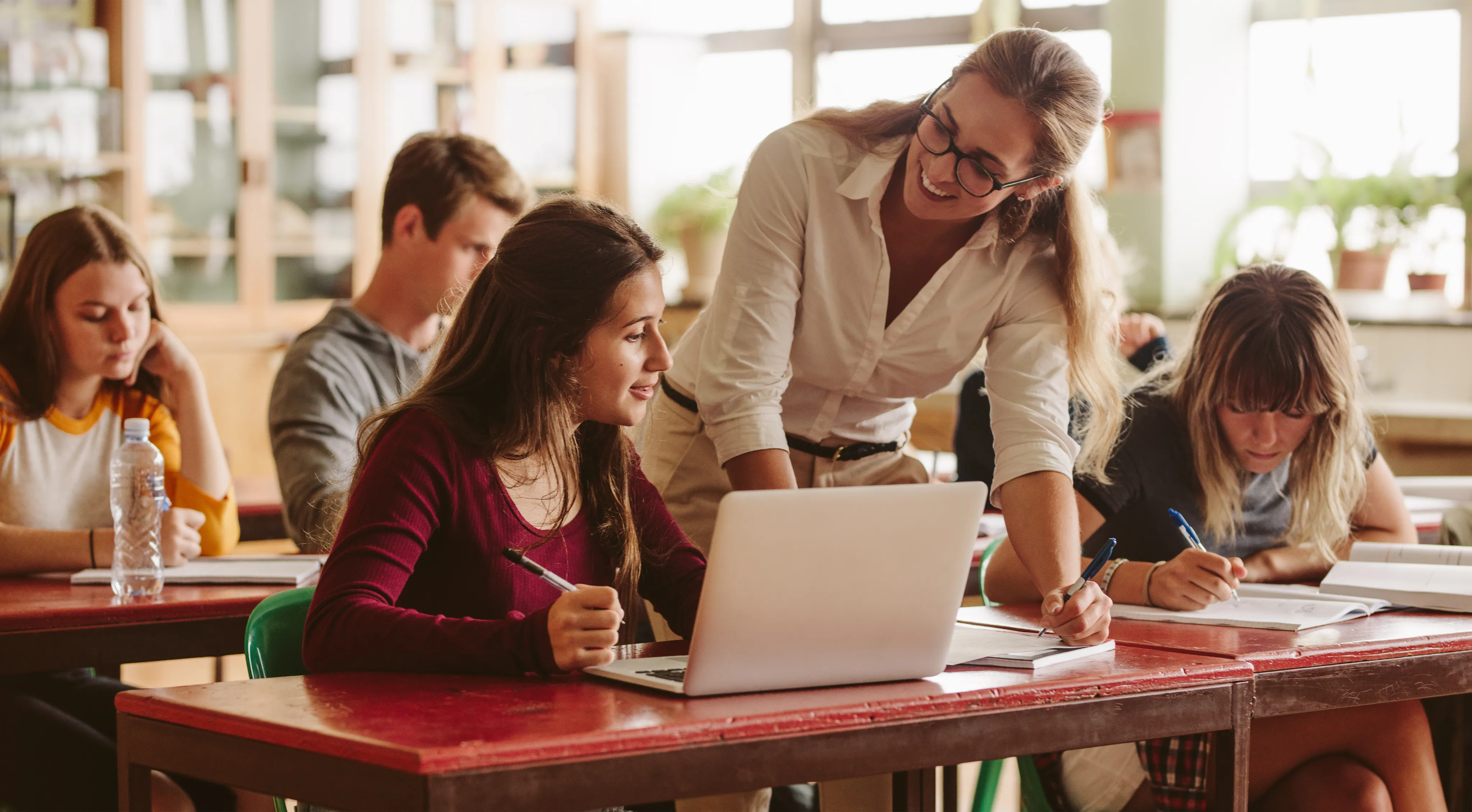 A teacher helps a student with a laptop in a classroom while other students work at their desks.