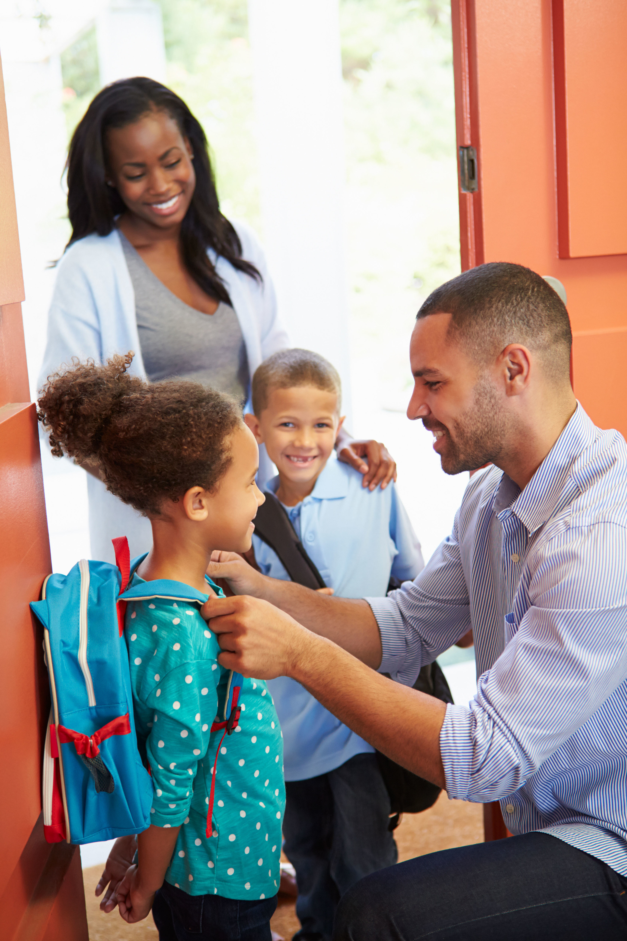 Smiling man helps a young girl with her backpack as a woman and boy stand nearby at the door.