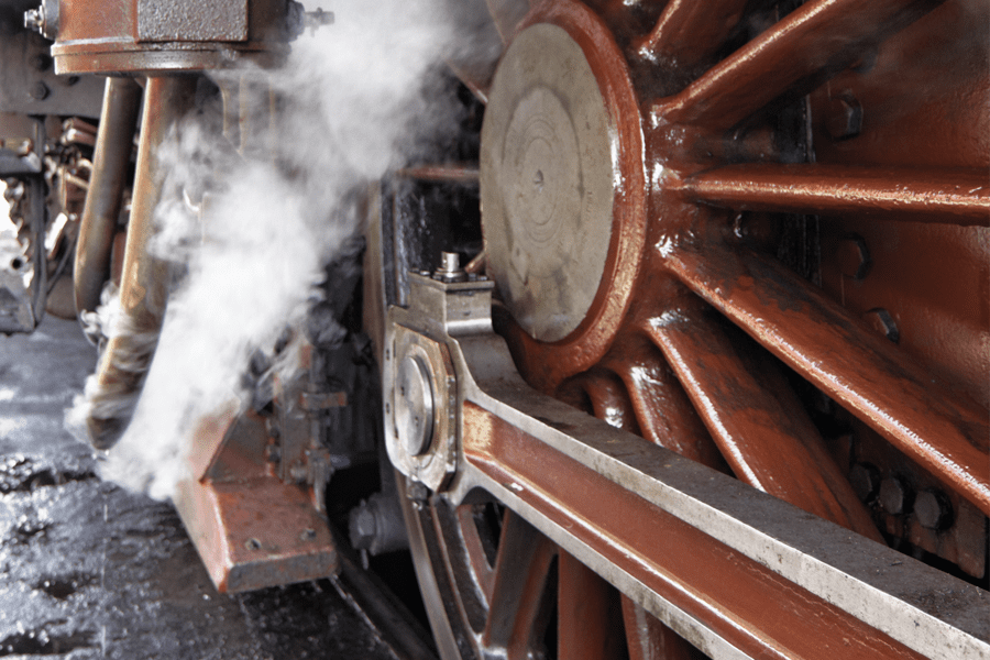 Close-up of a steam locomotive wheel with steam rising and metal parts visible.