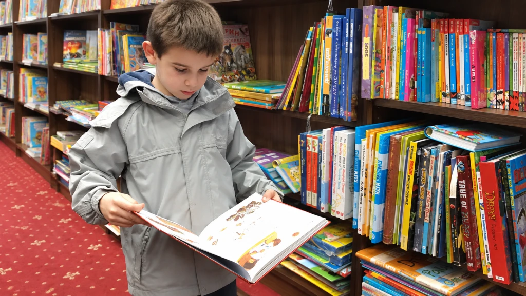 A boy in a gray jacket reads a book while standing by bookshelves in a library or bookstore.