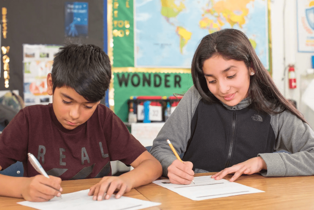 Two students sit at a desk writing on papers in a classroom.