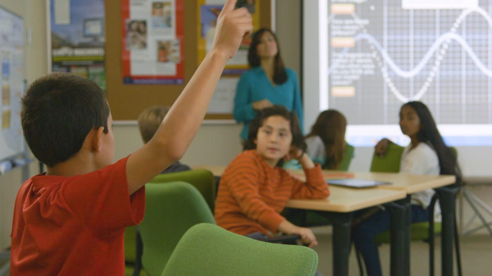 A student in a red shirt raises his hand in a classroom while others sit and a teacher stands by a screen.
