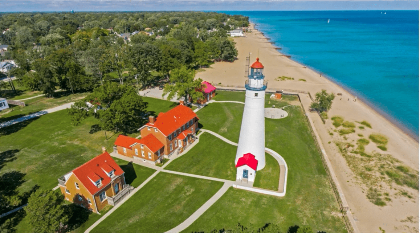 Aerial view of a white lighthouse and red-roofed buildings on a grassy shore next to a blue lake.