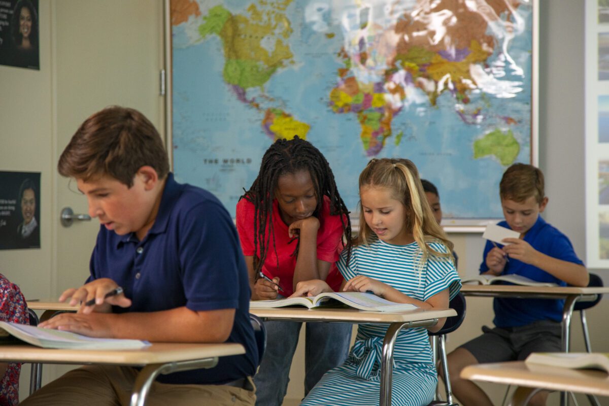 Five students sit at desks reading books in a classroom with a world map on the wall behind them.