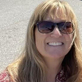 Woman with long blonde hair wearing sunglasses, smiling outdoors on a sunny day.