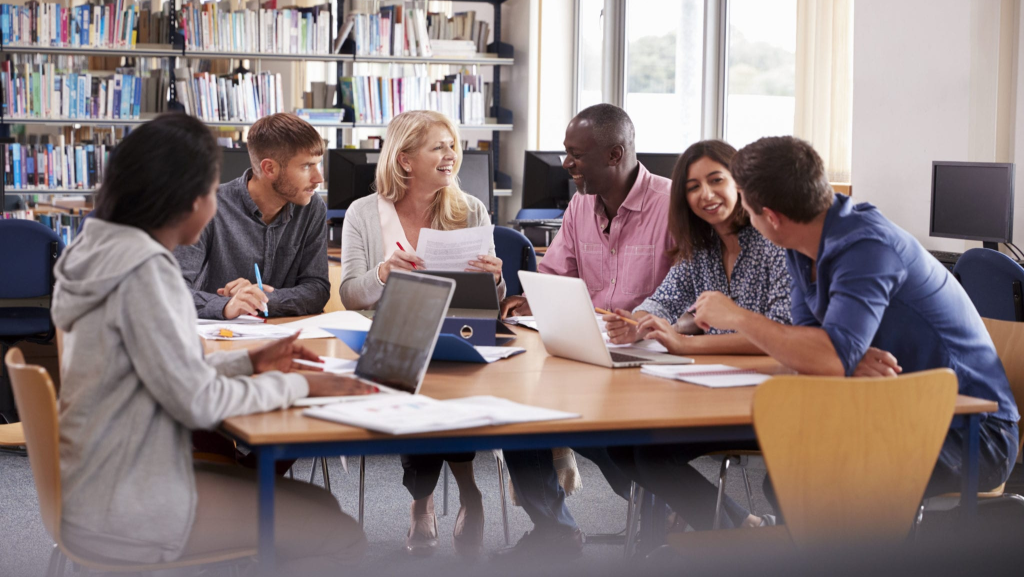 Six people sit at a library table, talking and smiling, with books and laptops in front of them.