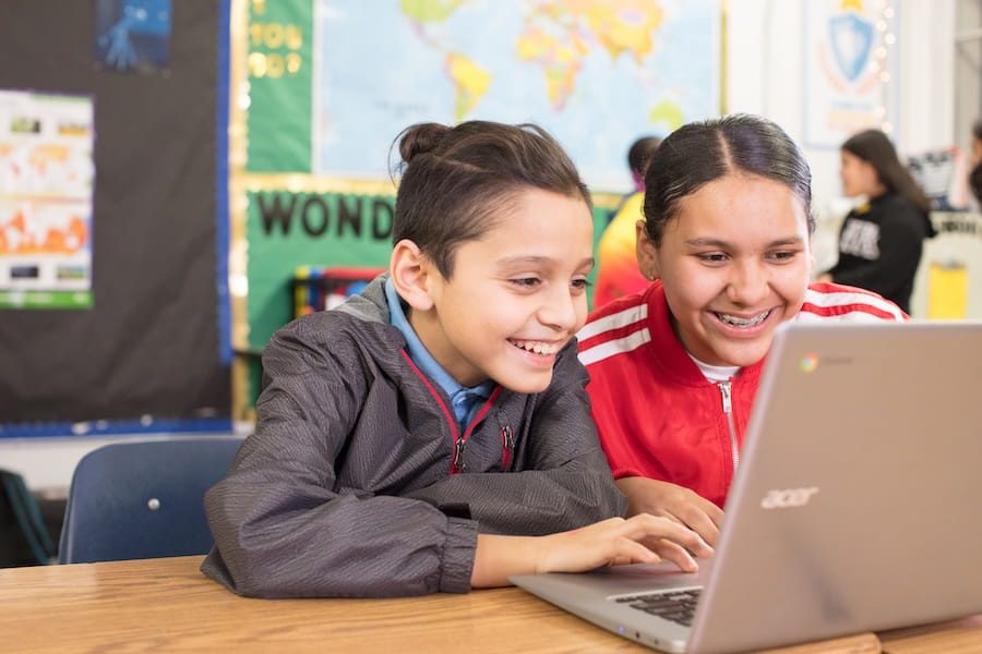 Two smiling kids sit at a desk, looking at a laptop together in a colorful classroom.