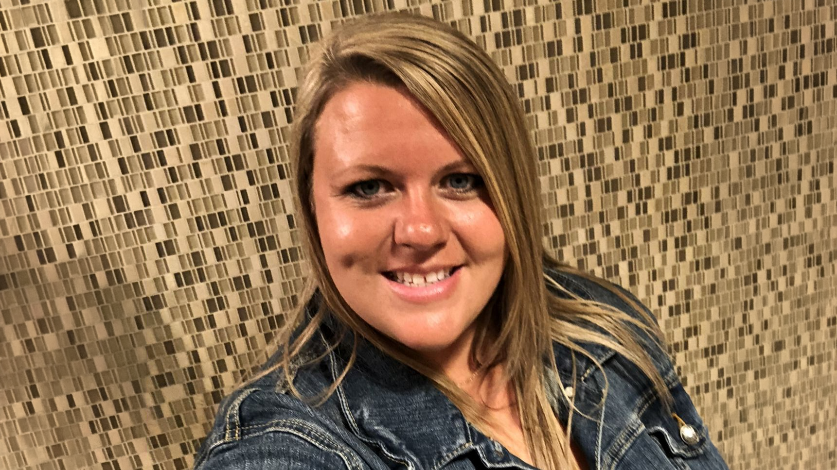 Woman with long blonde hair smiles in front of a mosaic tile wall, wearing a denim jacket.