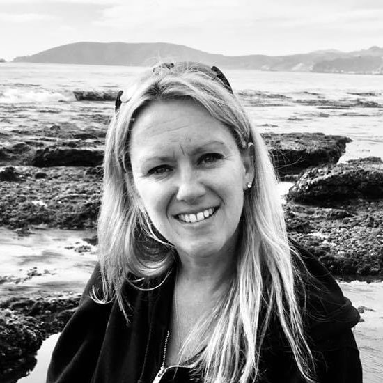 Woman with long blonde hair smiles at the camera; rocky shoreline and sea are visible in the background.
