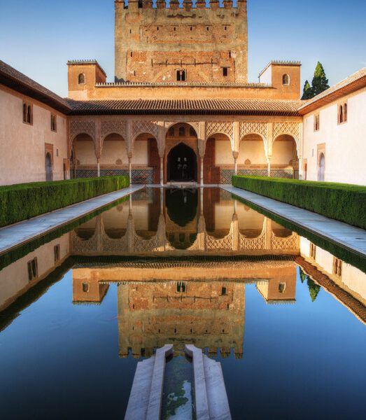 A palace courtyard with a large reflective pool, arched walkways, and ornate stonework under a blue sky.