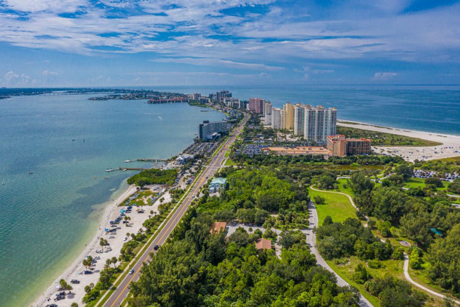 Aerial view of a coastal city with tall buildings, a beach, green parks, and a road along the shoreline.