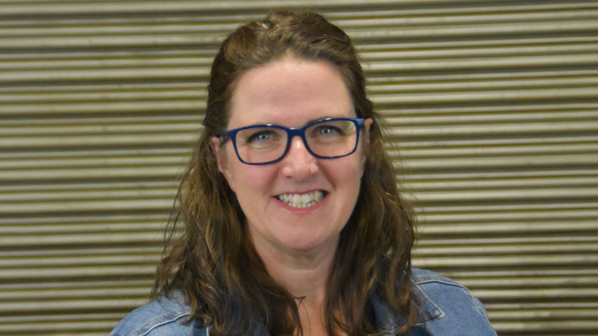 Woman with long brown hair and glasses smiling, wearing a denim jacket, in front of a striped metal background.