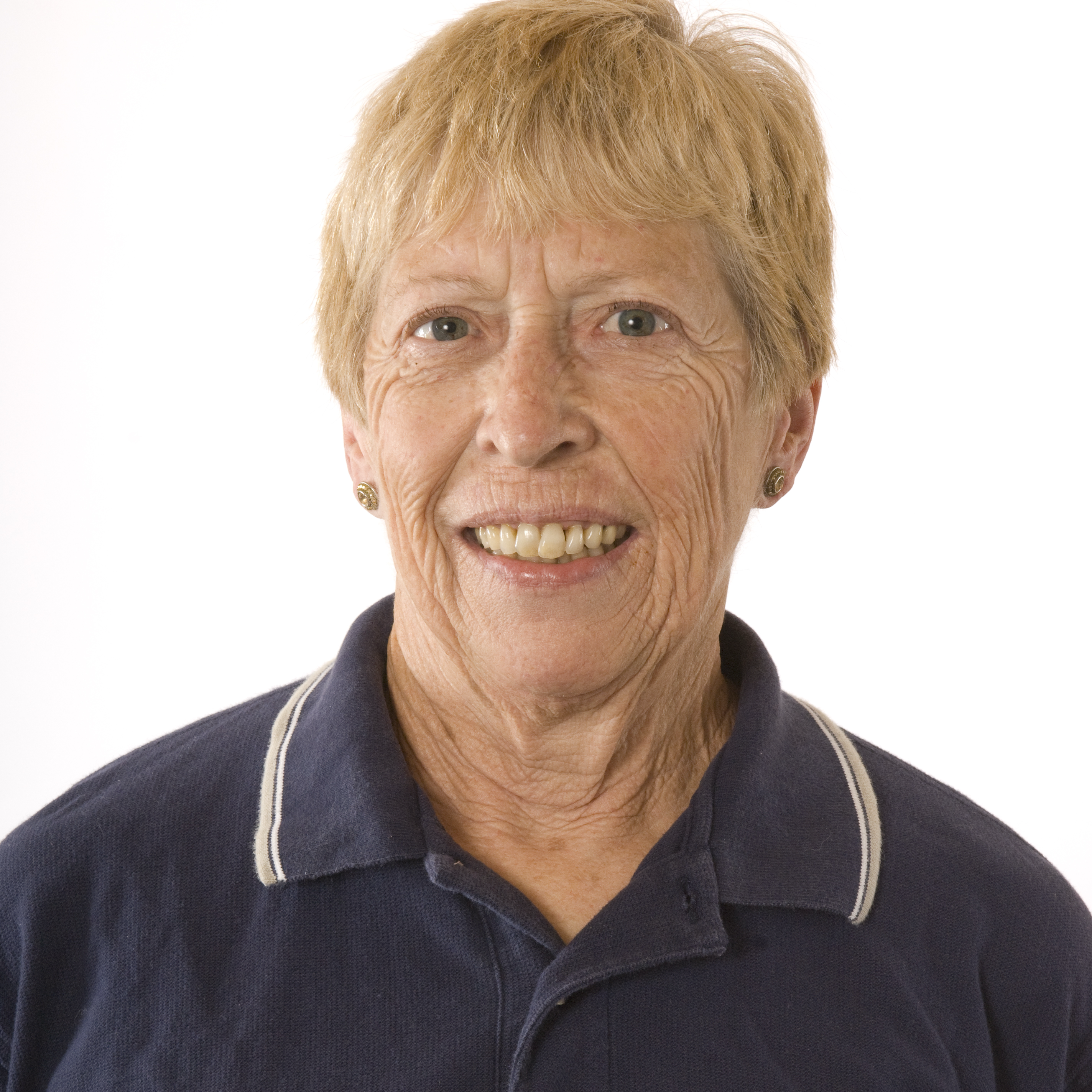 Smiling older woman with short blond hair wearing a navy collared shirt, facing the camera against a white background.