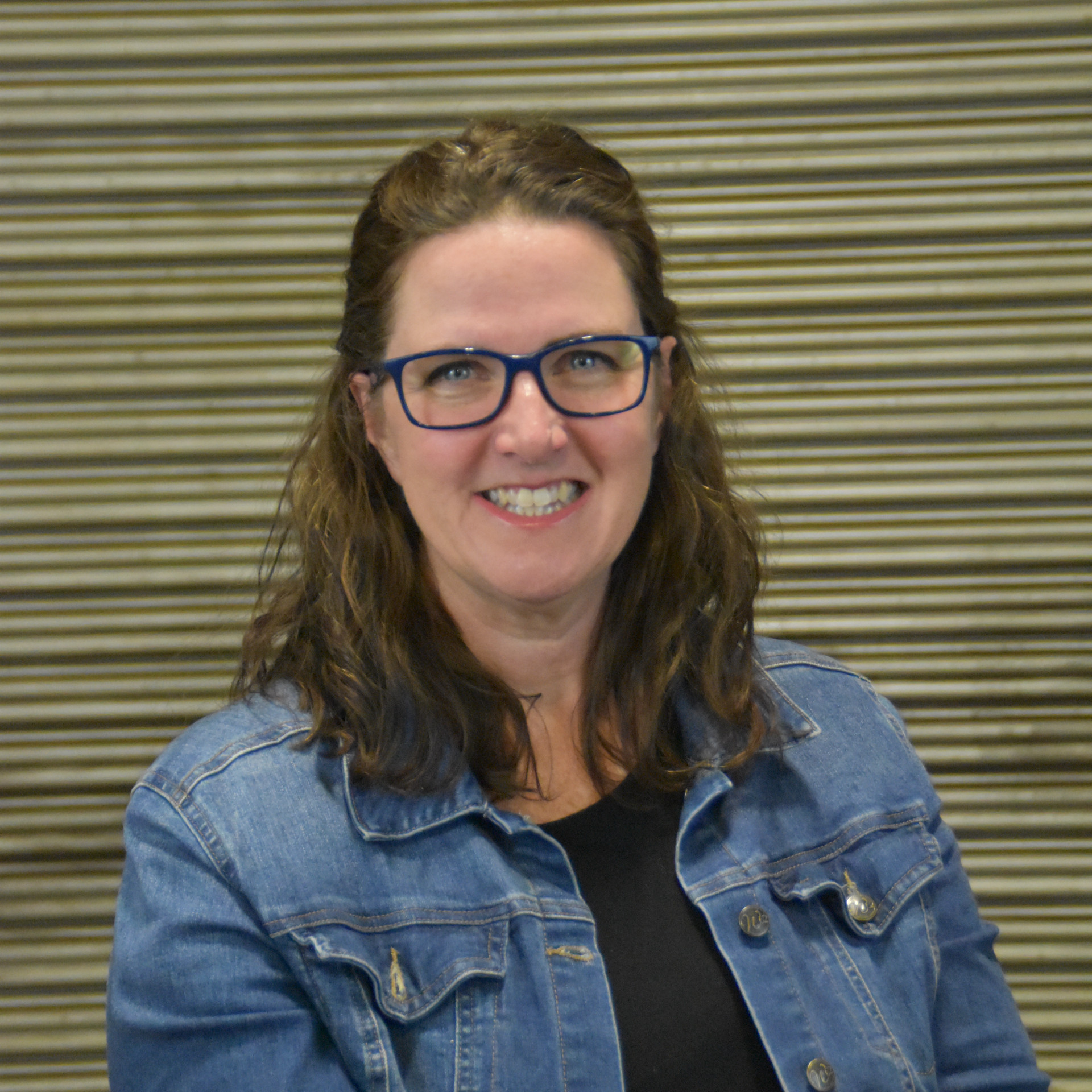 Woman with wavy brown hair and glasses, wearing a denim jacket, smiling in front of a striped background.