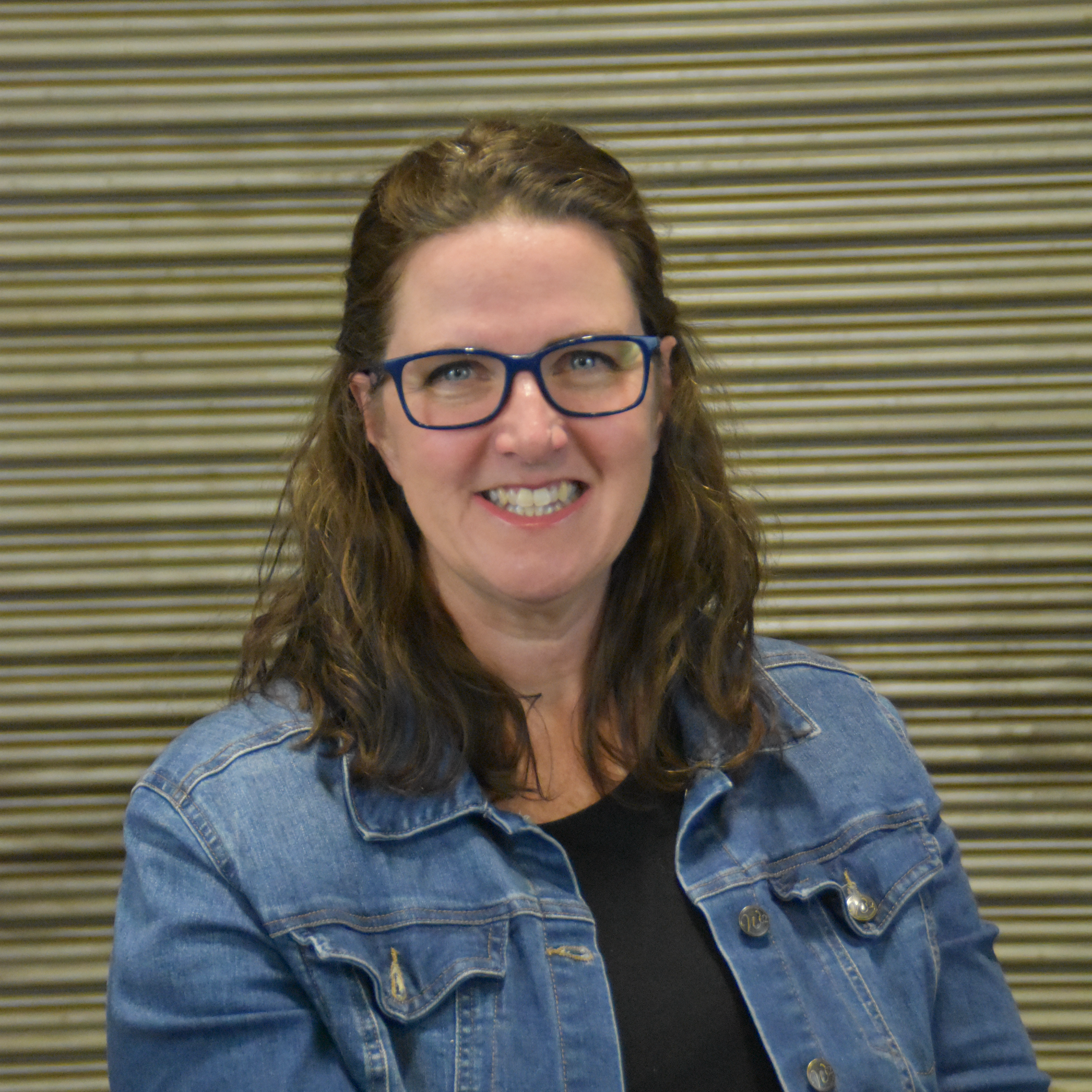 Woman with wavy brown hair and glasses, wearing a denim jacket, smiling in front of a striped background.