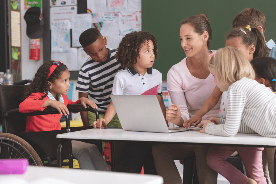 A teacher sits at a table with diverse children, including one in a wheelchair, looking together at a laptop.