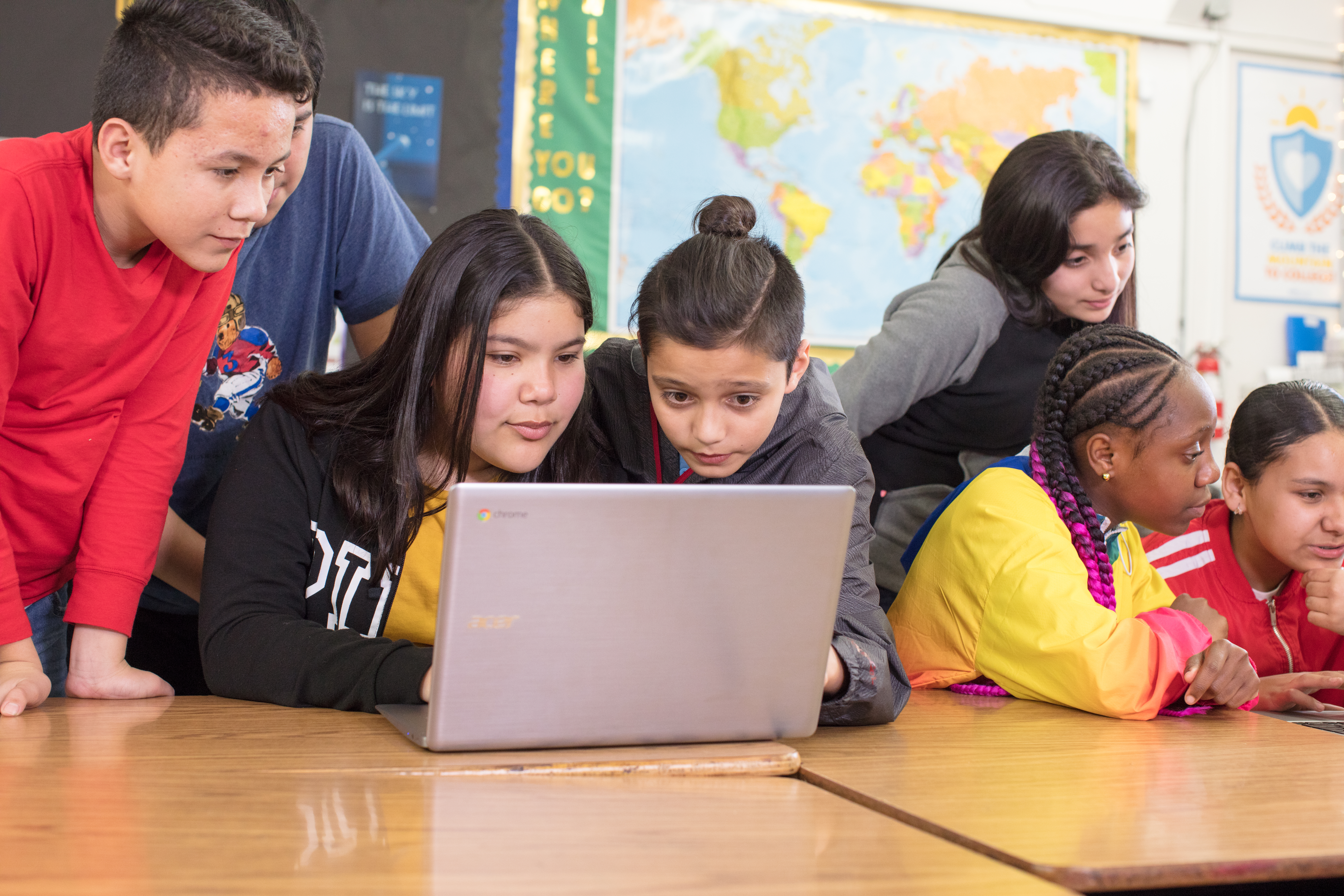 Group of middle school students gathered around a Chromebook in a classroom with a world map on the wall behind them.