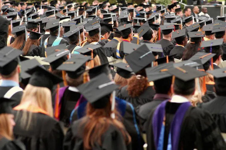 Graduates in black caps and gowns are seated outdoors at a commencement ceremony, facing the stage.