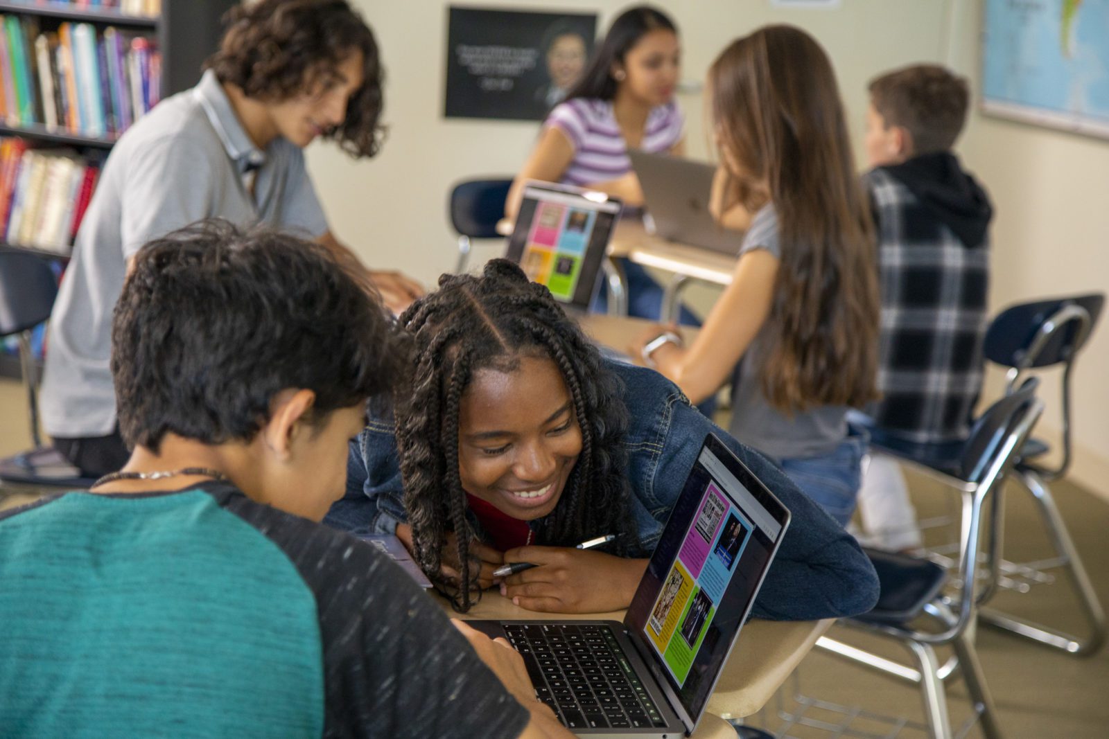 Students work together in a classroom, smiling and using laptops at their desks.