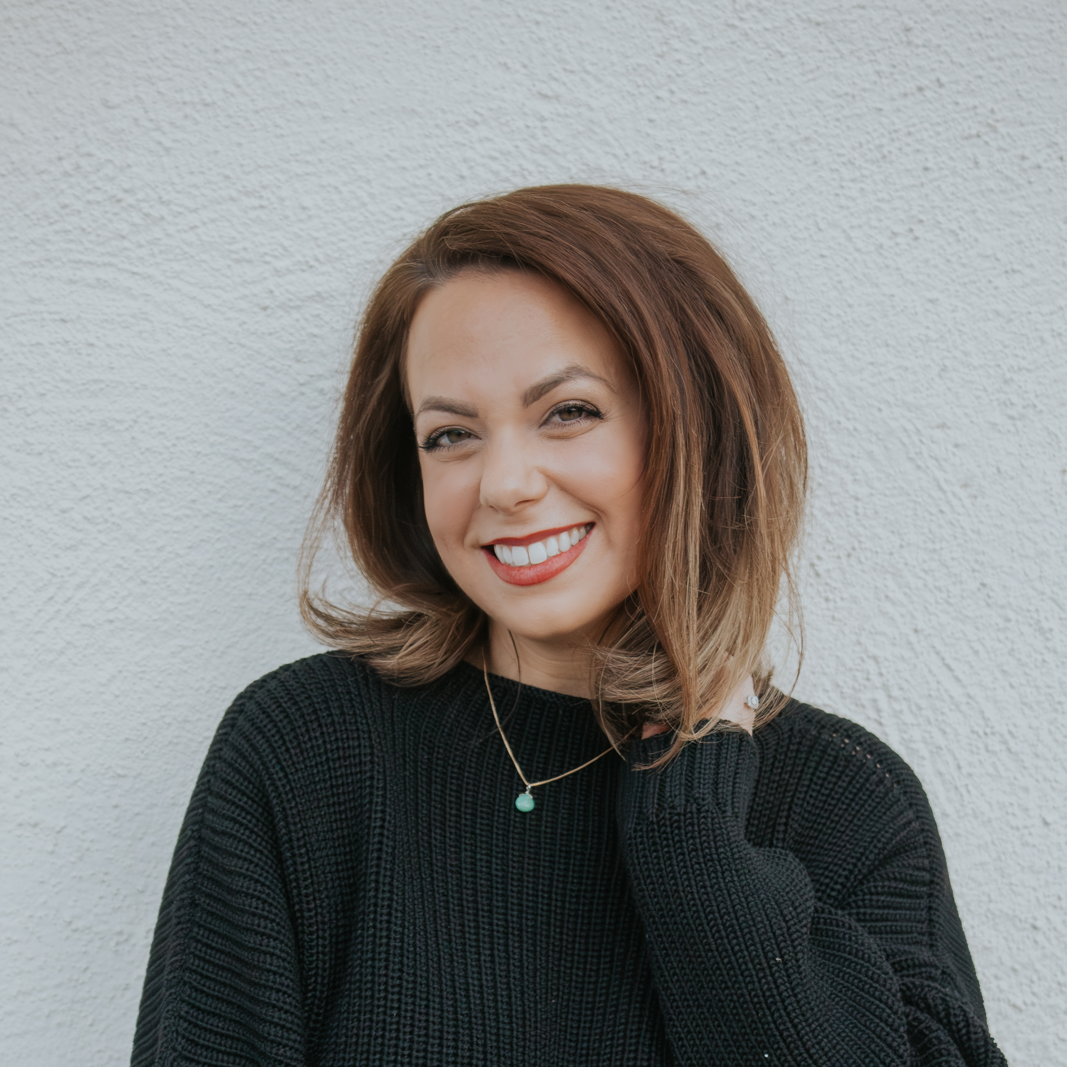 Woman with shoulder-length brown hair smiling, wearing a black sweater and a necklace, standing against a light wall.