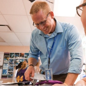 Smiling teacher in blue shirt and glasses helps a student at their desk in a bright classroom.