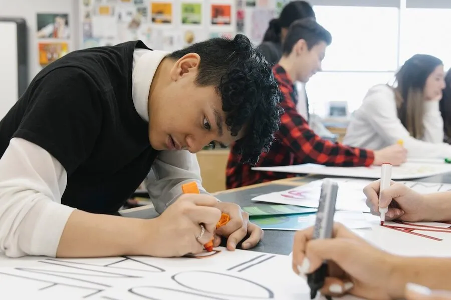 Student drawing with markers on poster board in a classroom, with others working in the background.