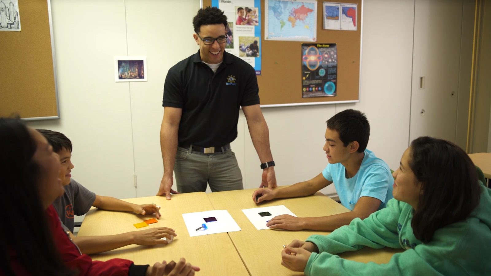 A teacher stands by a table, smiling at four students during a classroom activity with colored paper.