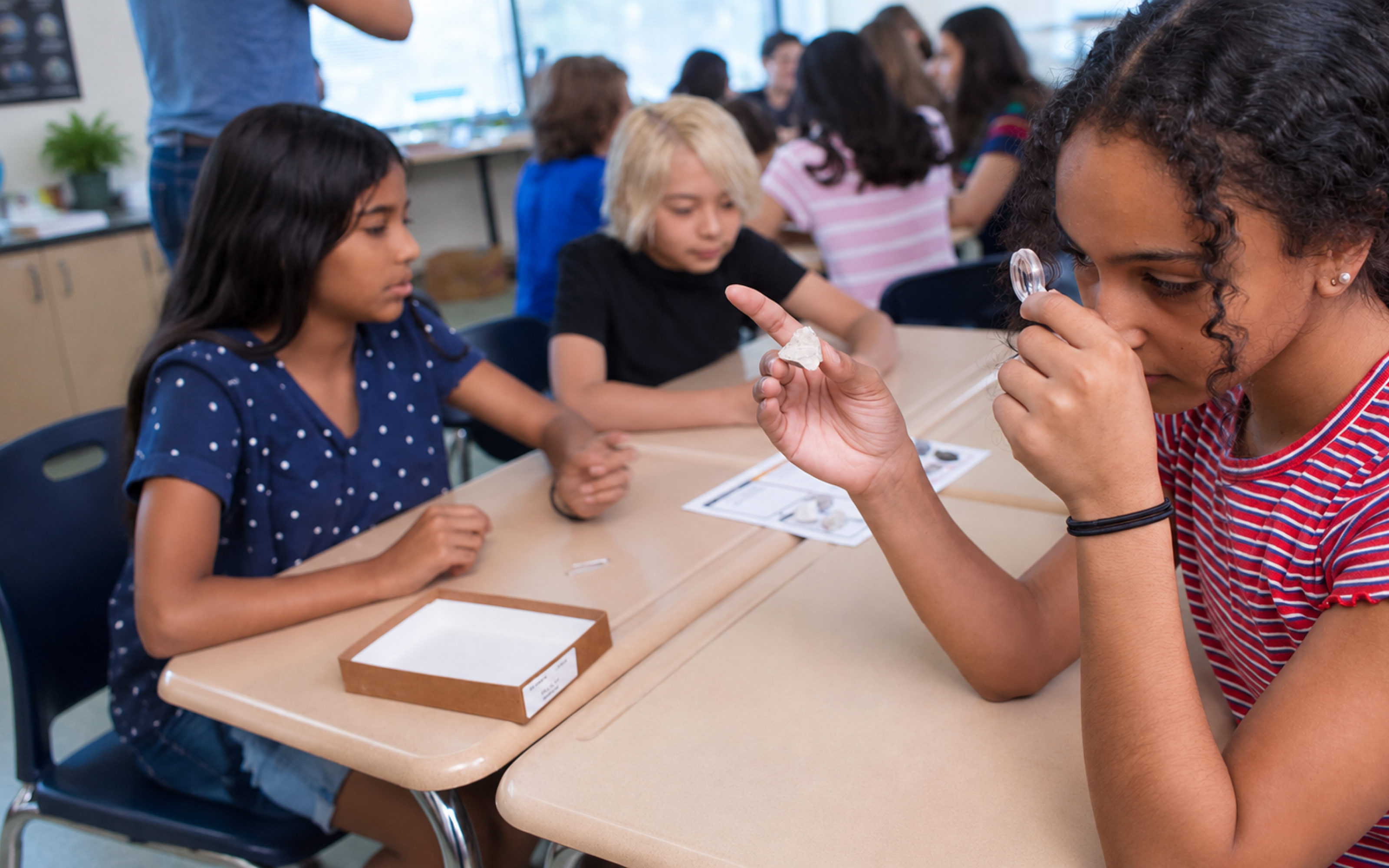 Three students examine rocks at a classroom table, one using a magnifying glass, others watching with interest.