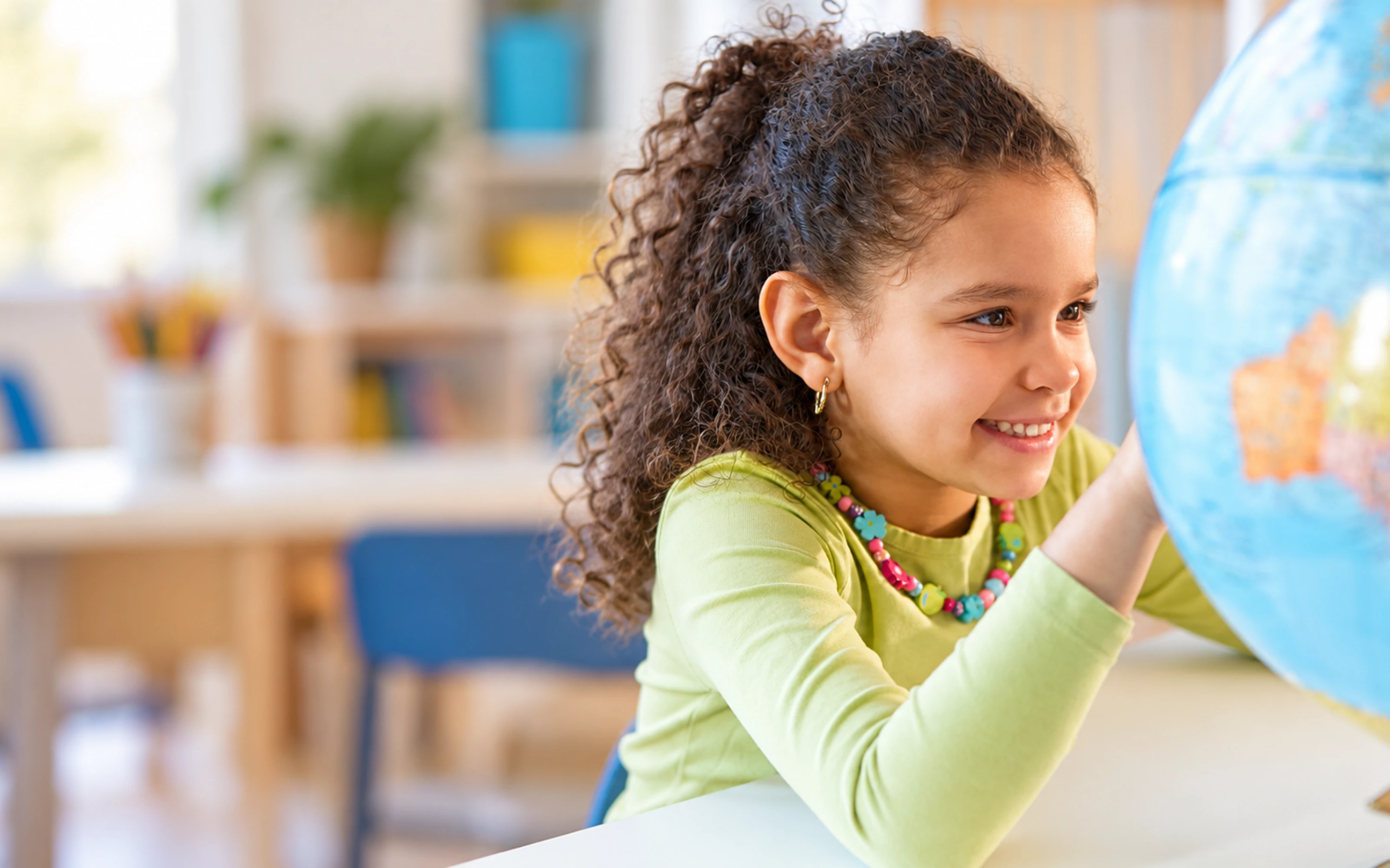 A young girl with curly hair and a colorful necklace smiles while looking at a globe in a classroom.