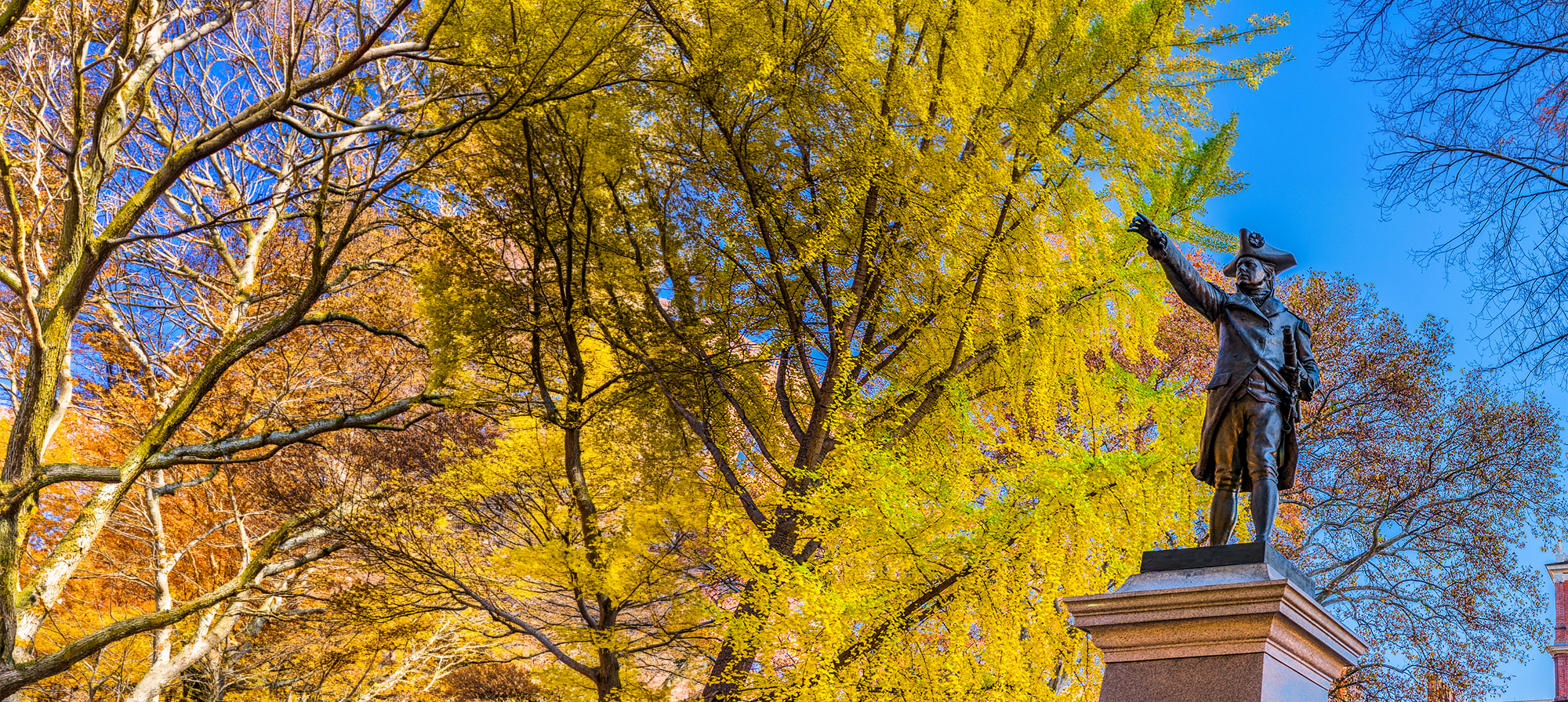 Statue on a pedestal in a park with colorful autumn trees and historic brick buildings in the background.