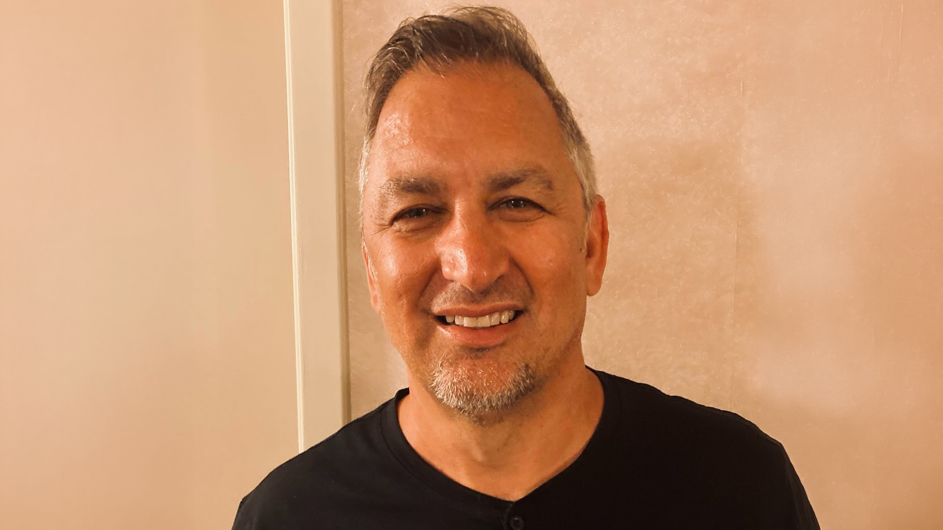 A man with short gray hair and a goatee smiles while wearing a black shirt, standing in front of a beige wall.