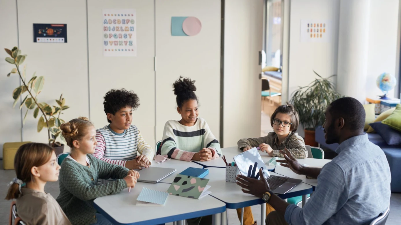A teacher talks to five children seated around a classroom table with notebooks and laptops.