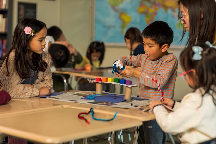 Four children and a teacher work on a colorful craft project at a classroom table with maps in the background.