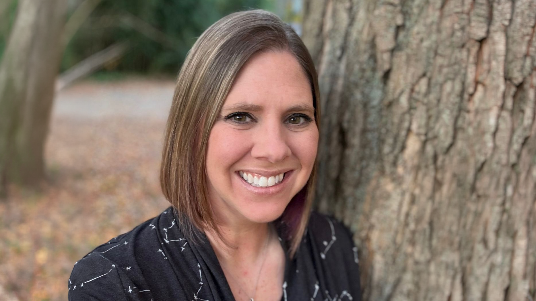 Woman with shoulder-length hair smiles beside a tree trunk, with autumn leaves and blurred greenery in the background.