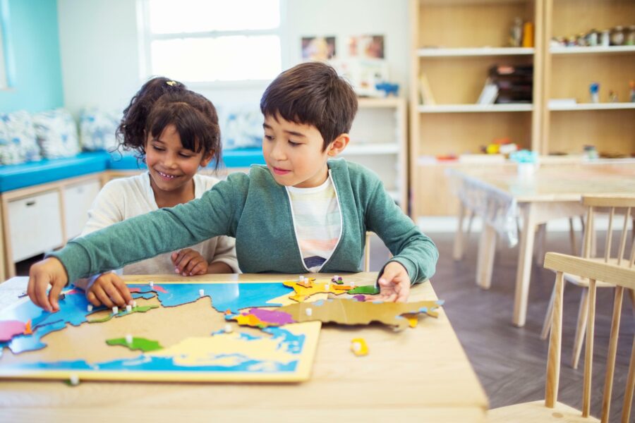 Two children sit at a table, working together on a colorful world map puzzle in a classroom.