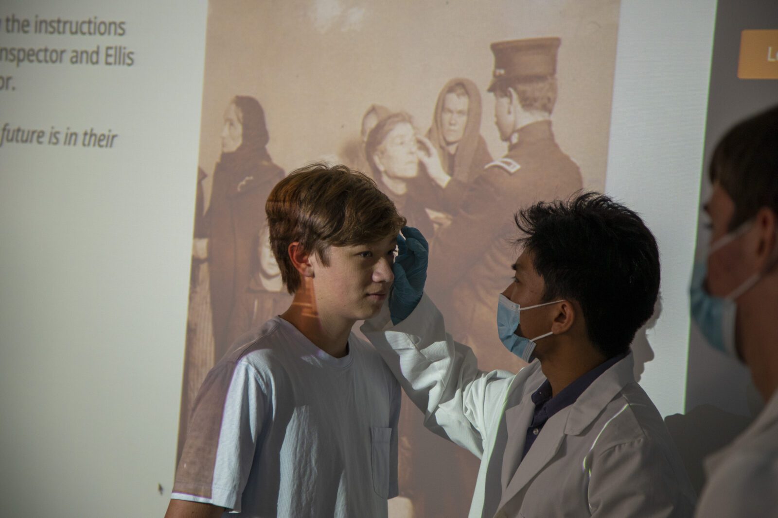 A person in a lab coat examines a boys eye, both wearing masks, with a historical photo projected behind them.