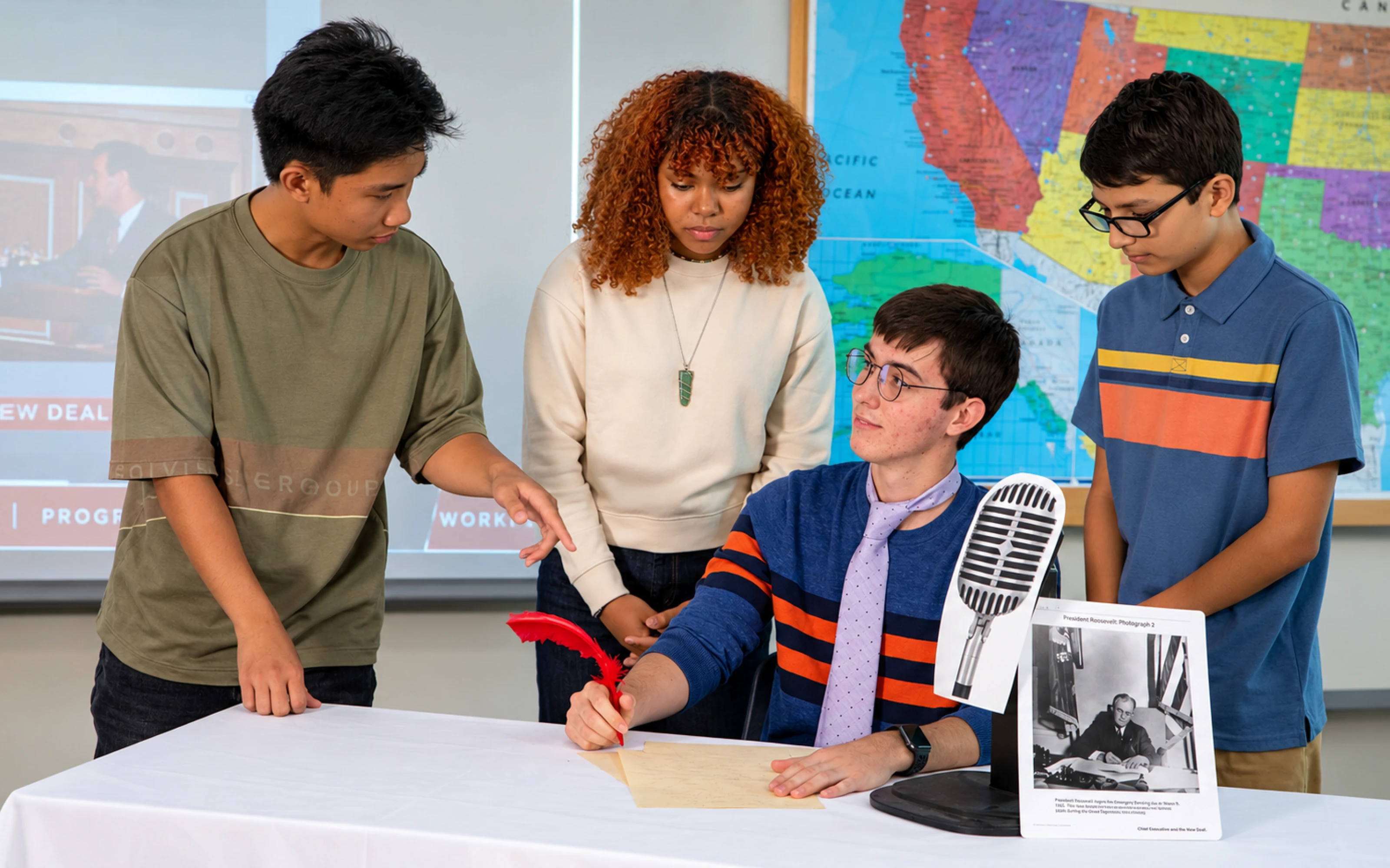 Four students role-play a historical scene with a desk, photo, vintage microphone, and a map in the background.