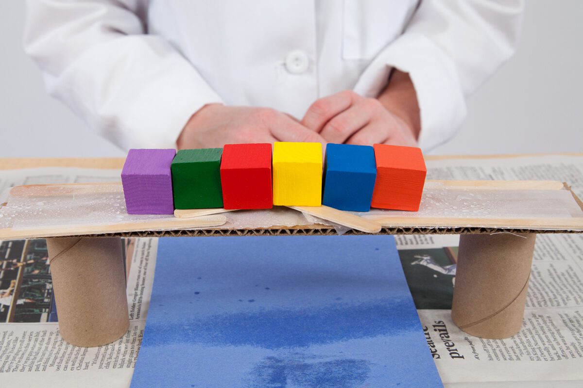 A person in a lab coat stands behind a bridge model with colorful blocks on top, built from cardboard and tubes.