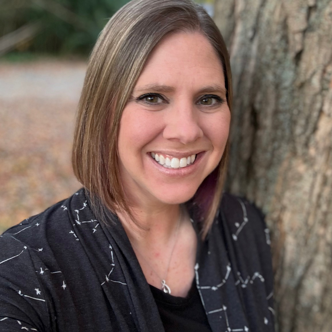 Woman with shoulder-length brown hair smiling, standing outdoors next to a tree, wearing a black patterned top.