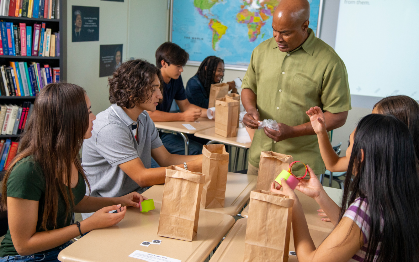 A teacher hands out paper bags to students in a classroom with books, a map, and a projector screen.