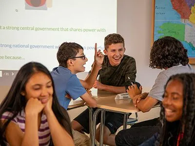 Five students sit at desks in a classroom, talking and laughing together in a group activity.