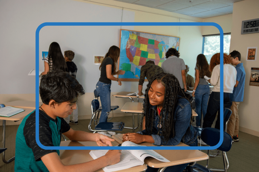 Two students study from a textbook at a desk while other students work in the background of a classroom.
