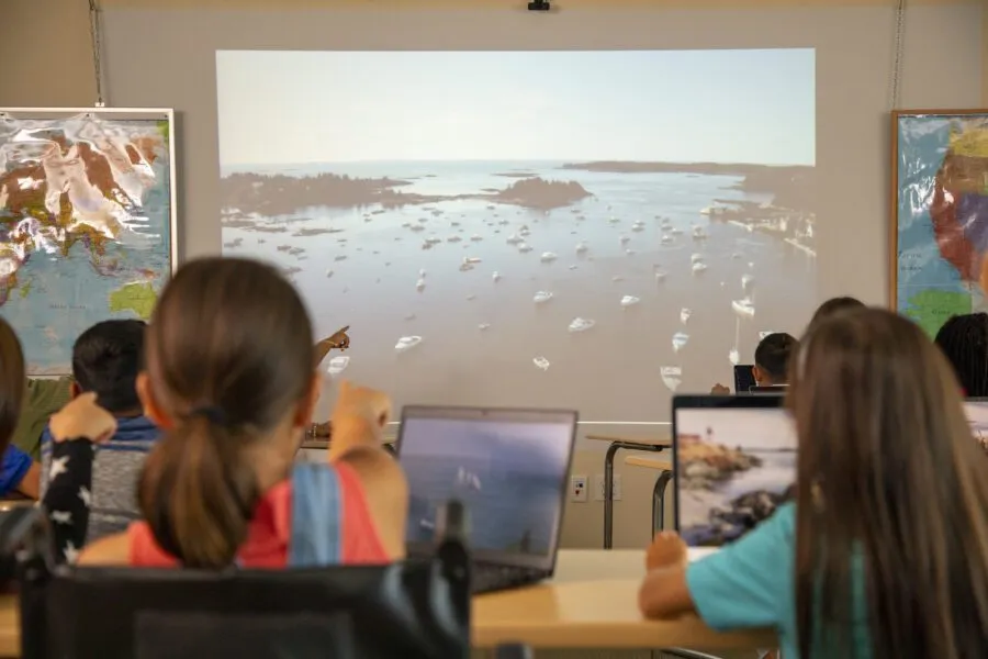 Students sitting at desks with laptops watch a projected image of a body of water with boats on it in a classroom.